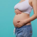Cropped photo of young pregnant female dressed in white sporty top and jeans holding raised hands on her belly while posing over blue background